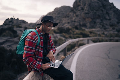 Male backpacker using laptop while sitting on railing at sunset