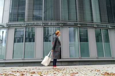 Man with tote bag standing by building on footpath