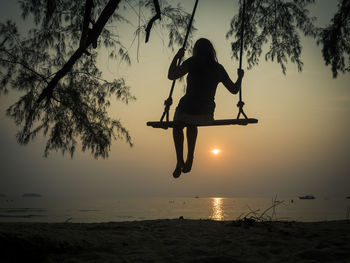 Rear view of girl sitting on swing at beach against sky during sunset