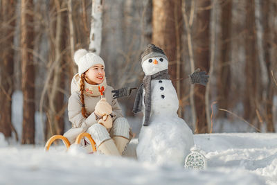 Teenage girl looking at snowman while sitting on sled in forest