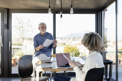 Senior man with paper smiling at woman while sitting on table at home