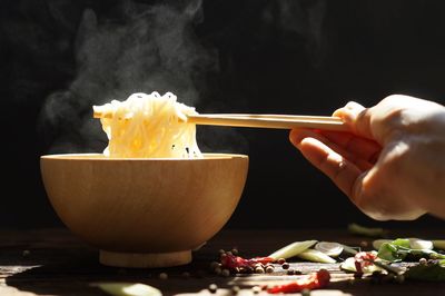 Hand picking up tasty steaming noodles with chopsticks in wood bowl and some spice on wooden table