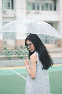 Side view of young woman holding umbrella standing on footpath against building