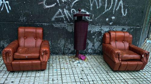 Dust bin amidst weathered sofas on sidewalk against wall