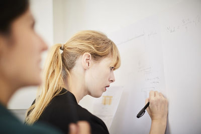 Side view of mid adult businesswoman writing on whiteboard with colleague in foreground at office