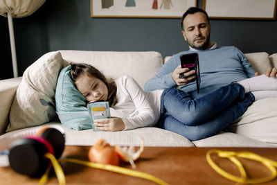 Father and daughter using mobile phones on couch in living room