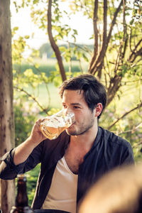 Young man looking away while drinking beer in glass against tree