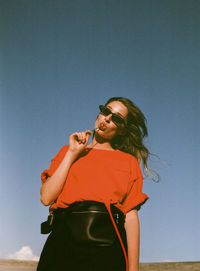 Low angle portrait of woman wearing sunglasses eating lollipop against clear sky