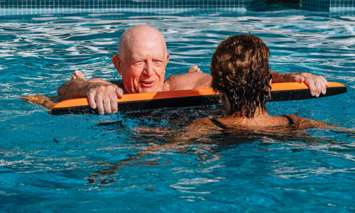 Happy senior husband leaning on aid kickboard while wife swimming in outdoor pool on sunny day