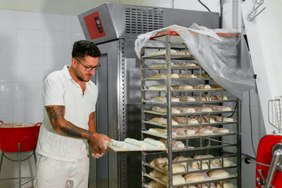 Young stylish tattooed male baker in casual clothes and eyeglasses putting loaves of raw dough on metal rack while baking bread in kitchen