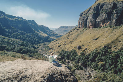 Rear view of woman looking at map while sitting on mountain against sky during sunny day
