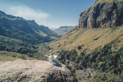 Rear view of woman looking at map while sitting on mountain against sky during sunny day