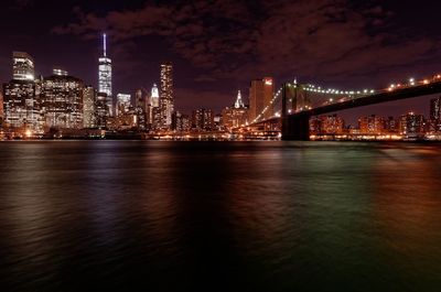 Illuminated bridge over river by buildings against sky at night