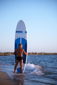 Rear view of shirtless man holding surfboard standing at shore against clear sky