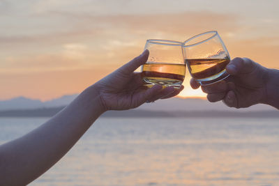 Cropped hands of couple toasting whiskey against sky during sunset