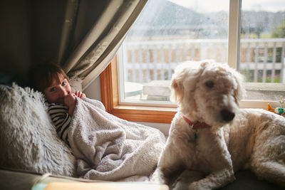 Portrait of girl with dog sitting on bed by window at home