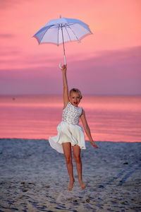 Full length of surprised girl flying while holding umbrella at beach against dramatic sky during sunset