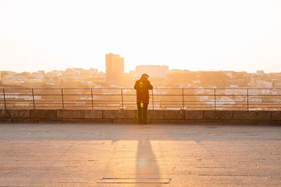 Rear view of man standing road against cityscape against sky during sunrise