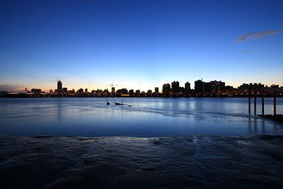 Silhouette buildings by river against clear sky at sunset