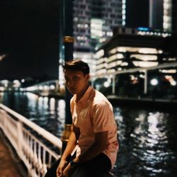 Young man sitting on railing by river in city at night