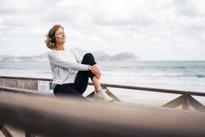 Mature woman wearing wireless headphones sitting on railing at beach
