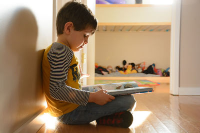 Side view of boy reading book while sitting on floor at home