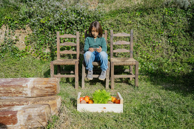 Boy sitting in garden with vegetable box, playing games on his smartphone