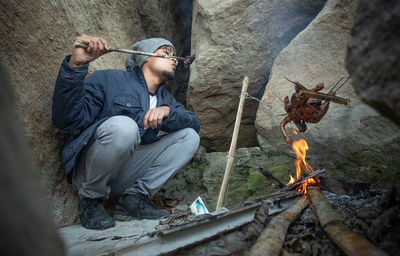 Man roasting meat over fire while sitting by rocks