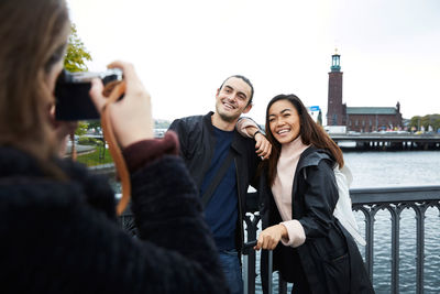 Woman photographing smiling friends standing on bridge by railing in city against clear sky