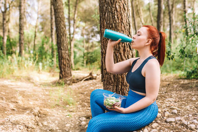 Woman drinking water through bottle sitting by tree