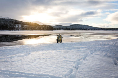 Rear view of man crouching on lakeshore against cloudy sky during winter