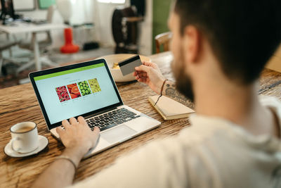 Crop unrecognizable male making purchase with plastic card for order during online shopping via laptop