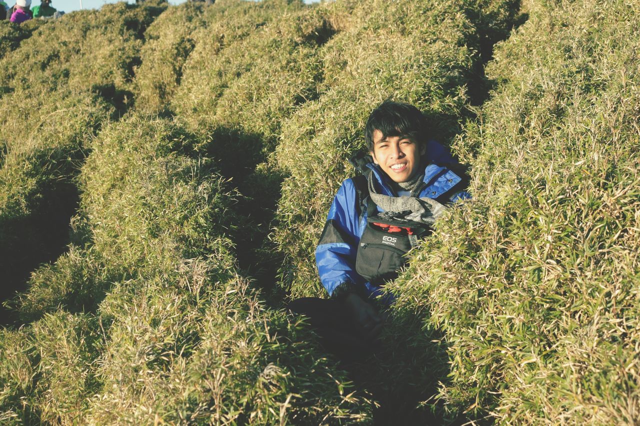 lifestyles, leisure activity, casual clothing, grass, person, growth, field, green color, plant, portrait, looking at camera, smiling, young adult, standing, nature, high angle view, day, grassy