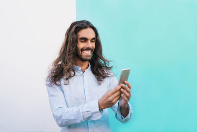 Smiling man with long hair using smart phone against wall