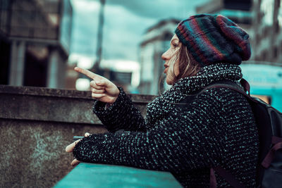 Woman in warm clothing pointing by retaining wall in city