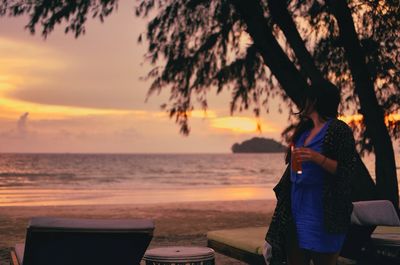 Young woman with drink standing at beach during sunset