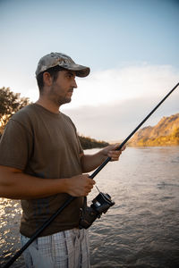 Man holding fishing rod standing by river standing against sky