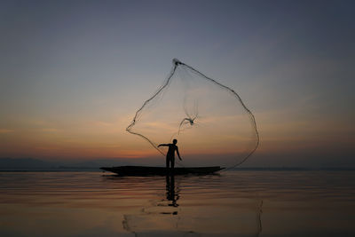 Silhouette fisherman standing by sea against sky during sunset