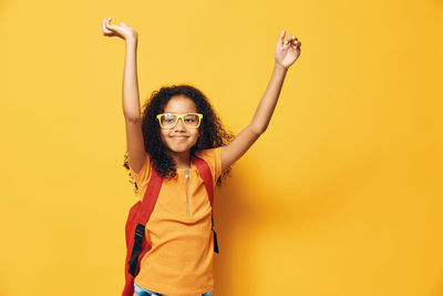 Portrait of young woman with arms raised standing against yellow background