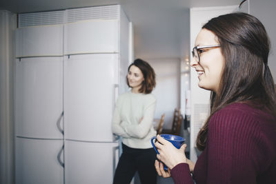 Side view of happy woman with coffee cup standing against female friend in dorm