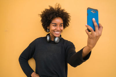 Smiling young woman taking selfie standing against wall