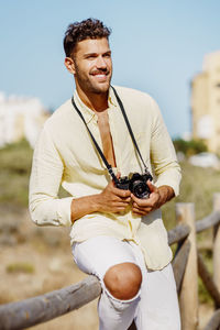 Smiling young man holding camera standing by railing against sky