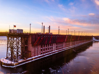 Factory by river against sky during sunset