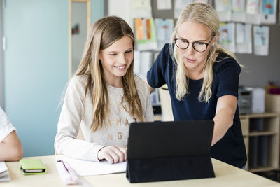 Teacher assisting happy girl in using digital tablet at school