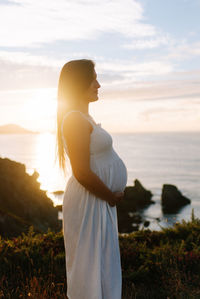 Side view of woman standing at beach against sky during sunset