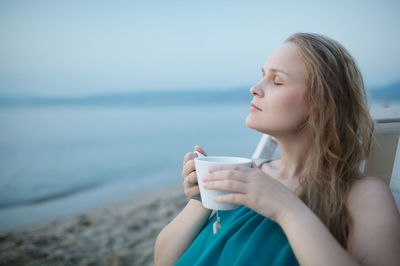 Woman with eyes closed holding coffee while sitting on chair by sea against sky