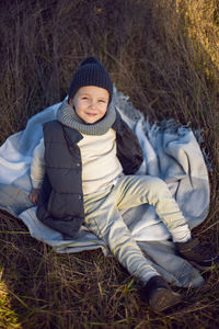 Boy in warm clothes sit on the bedspread along a path on a field with dried grass in autumn