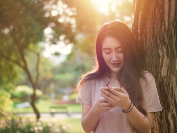 Beautiful young woman using mobile phone against tree