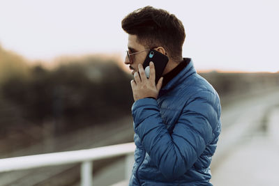 Young man in blue jacket talking on smart phone during sunset