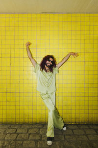 Playful man legs crossed at knee and arms raised standing near yellow wall